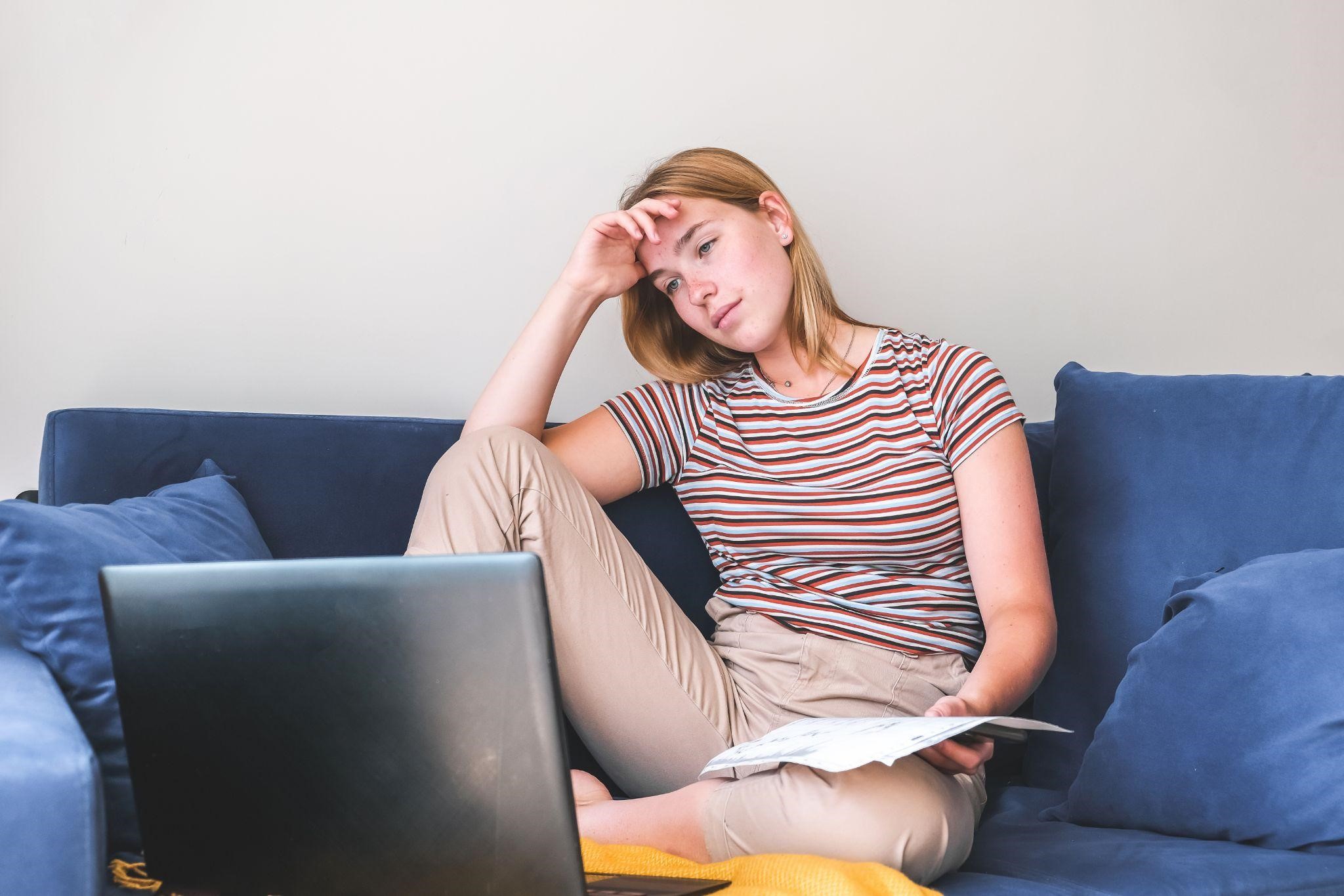 A girl, looking tired, works on her computer while holding newspapers and sitting on the couch